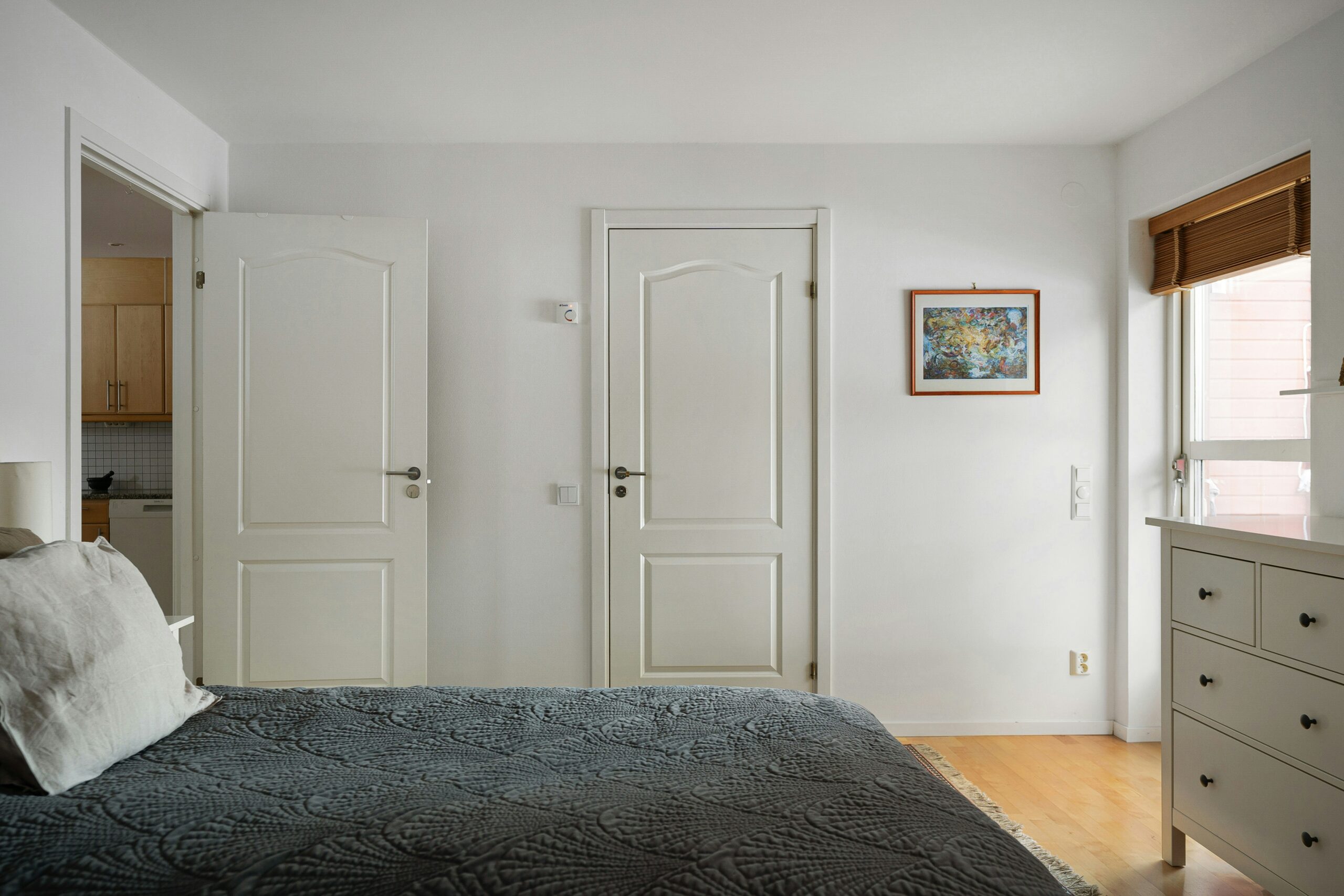 A photograph of a bedroom with the door open, revealing a kitchen in the next room. A double bed with a gray bedspread takes up most of the room, with a dresser and window on the opposite wall. The walls are white.