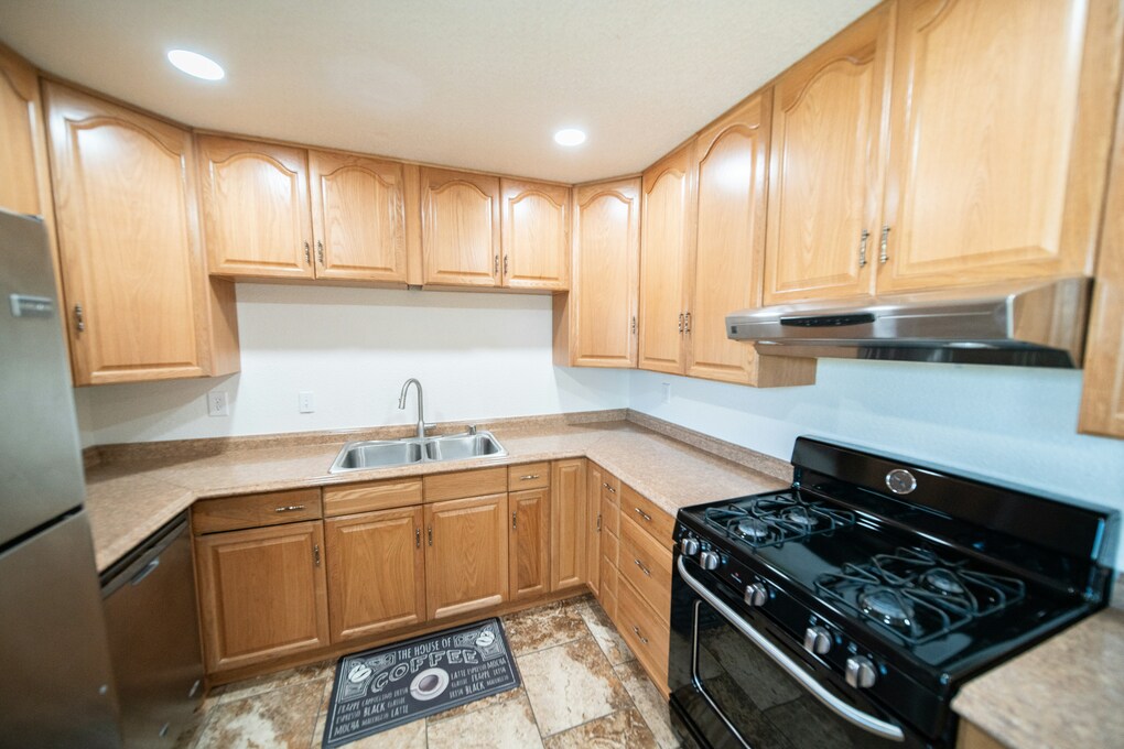 A photograph of a kitchen with natural colored wooden cabinets, black stove, tan laminate countertops and brown tile floor.