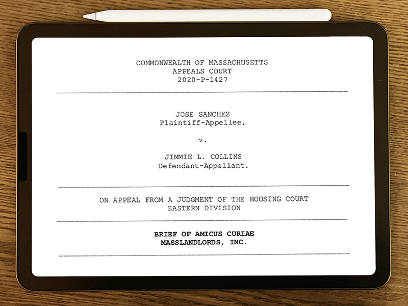 An ipad with an attached white Apple pencil sits on top of a dark-grain wooden table. On the iPad’s screen, the first page of an amicus brief is visible, black text on a white background reading “Commonwealth of Massachusetts Appeals Court 2020-P-1427, Jose Sanchez, Plaintiff-Appellee, v. Jimmie L. Collins Defendant-Appellee, on appeal from a judgment of the housing court eastern division, Brief of Amicus Curiae, MassLandlords, Inc.”