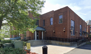 A 2-story brick building surrounded by trees and a small brick courtyard. A parking lot can be partially seen in the foreground.