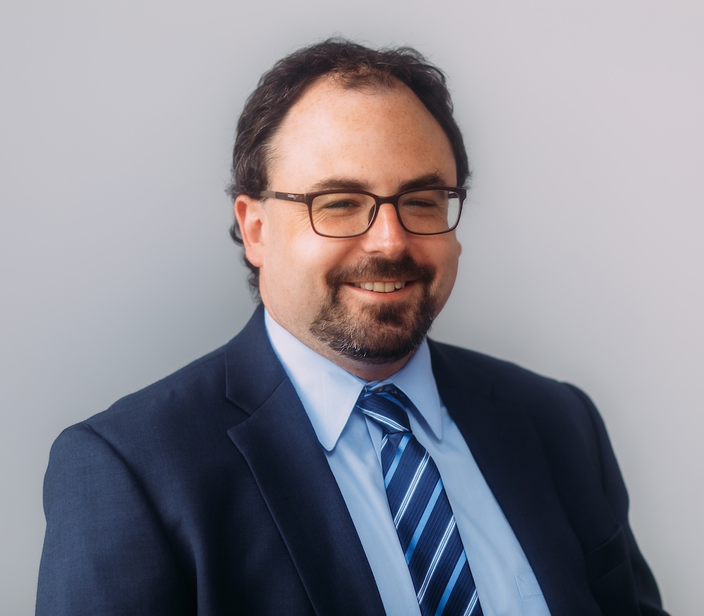 Professional headshot of attorney Adam Sherwin, in navy suit and patterned tie. Attorney Sherwin wears glasses and a goatee and moustache.