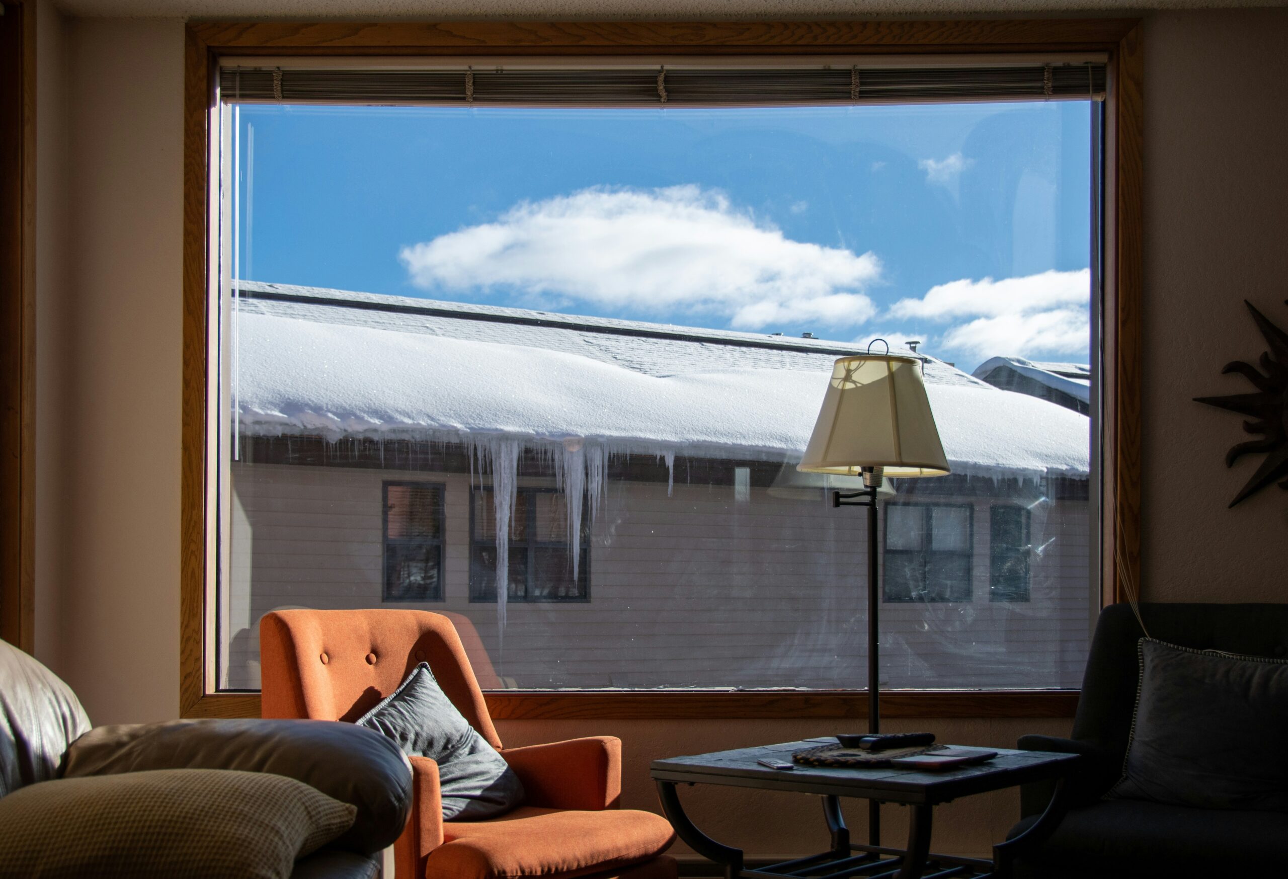 A photograph of a minimalist living room with big picture window looks out onto another house which has lots of snow and icicles on the lower portion of the roof, but no snow on the peak.