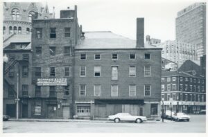 Black and white photo of the Jenney building in Boston, circa 1970s-80s, with boarded up windows and storefronts, with State Street Bank and other downtown Boston buildings in the background, and a few 1970s-era cars parked along the curbs.
