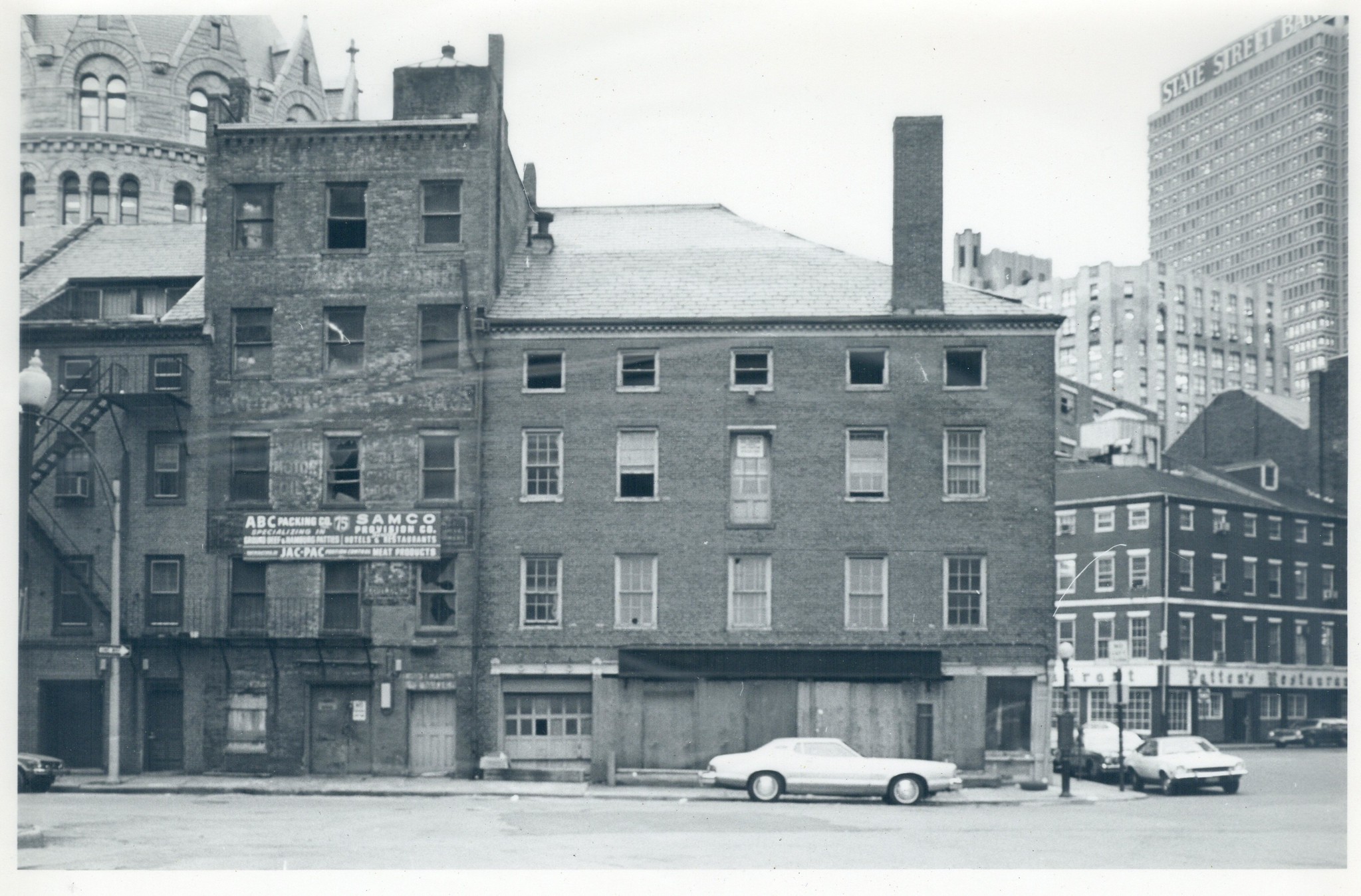 Black and white photo of the Jenney building in Boston, circa 1970s-80s, with boarded up windows and storefronts, with State Street Bank and other downtown Boston buildings in the background, and a few 1970s-era cars parked along the curbs.