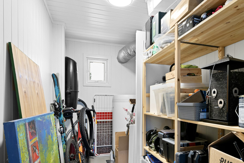 A brightly lit storage area with wood paneling and wood shelves contains a painting, a table, skis, a bicycle, some wire metal shelves, two cardbox boxes and personal effects.