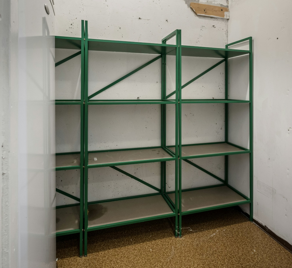 A storage area with empty metal-framed, fiberboard shelves sits empty. A cork floor, piece of wood screwed to the wall and stain on the shelf suggest this is a budget storage area.