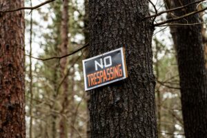 [Alt: A sign with “No Trespassing” in bold letters on a black background is nailed to a tree in a wooded lot.]