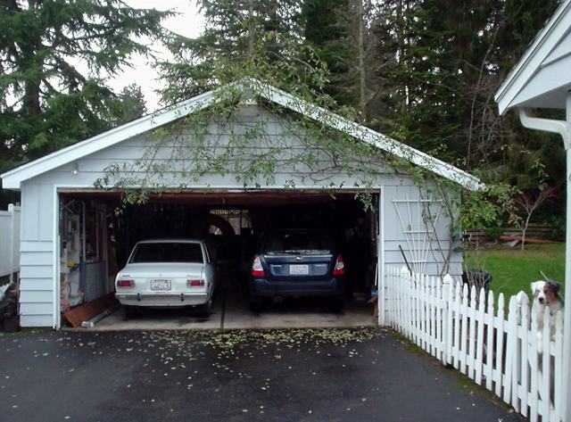 A wood-frame, two-car garage sits at the end of a driveway. The driveway is lined by a white picket fence. Inside the garage are an older sedan and a newer SUV.