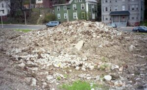 photo of an urban vacant lot covered in a pile of gravel and large rocks, across the street from multifamily houses.