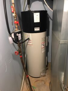 Photo of a black and white heat pump water heater in a basement corner, with water pipes, hoses and a meter affixed to it, and a large air duct running alongside.
