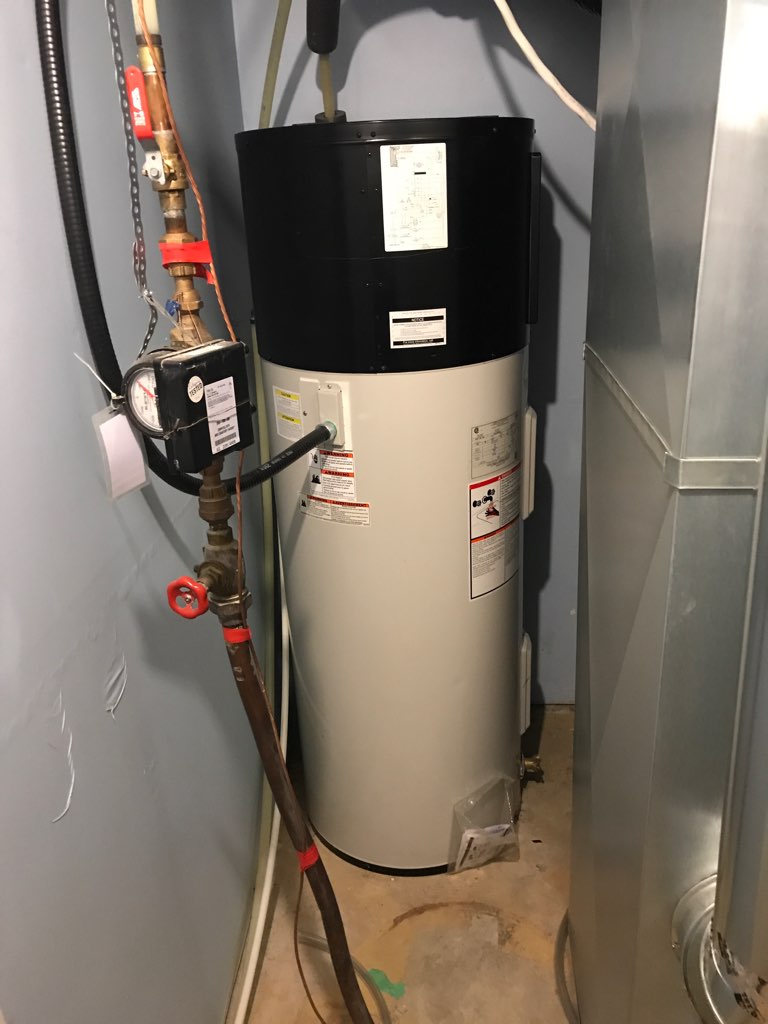 Photo of a black and white heat pump water heater in a basement corner, with water pipes, hoses and a meter affixed to it, and a large air duct running alongside.