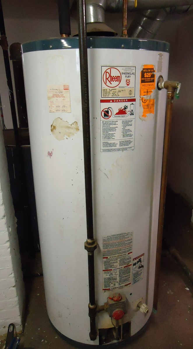 Photo of a gas-powered storage tank water heater, white, with pipes running up the length of the unit and some brand and technical decals affixed to the outside, in a basement. 