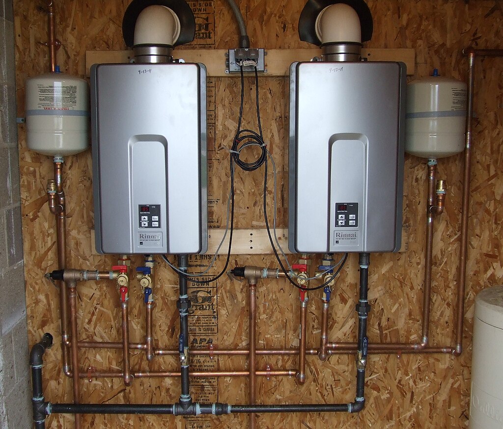 Photo of two gas-powered tankless water heaters with gray covers, affixed to a large, composite panel board with copper gas lines running into and out of each unit, with a small, white overflow tank beside each unit and electric wires dangling from each unit, plugged into an outlet.