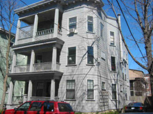 A photograph of a gray triple-decker house with a red SUV parked in front