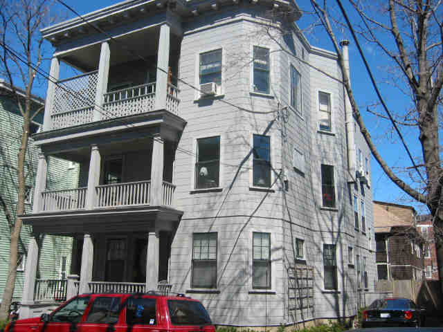 A photograph of a gray triple-decker house with a red SUV parked in front
