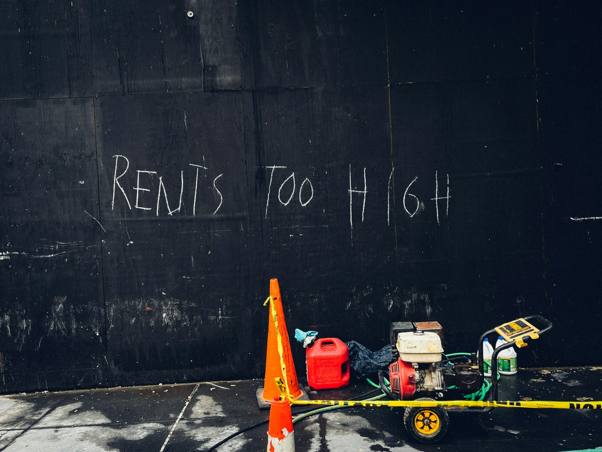 A plywood wall painted black sits along a sidewalk. Power tools and gas cans sit in front of it, blocked off with caution tape. Someone has written “rents too high” in white on the wall.