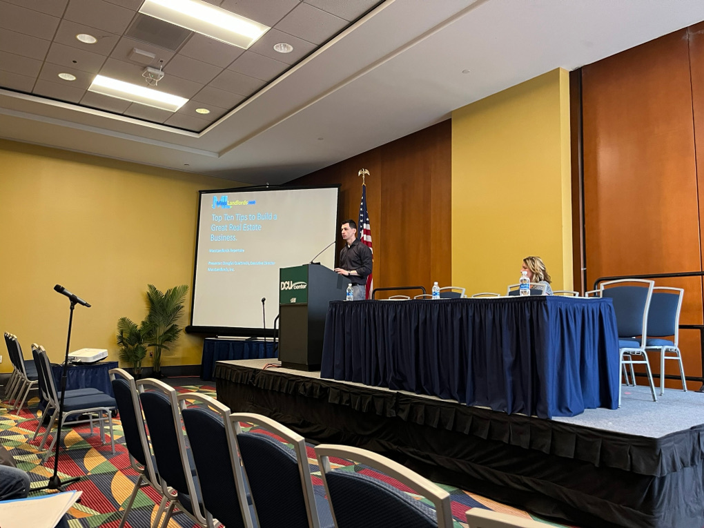 In a DCU ballroom, Doug Quattrochi stands on a raised platform at a podium. Behind him is the title slide of this presentation and the American flag. He wears a watch and a collared shirt. The front row is empty but there are attendees seated in the second row.