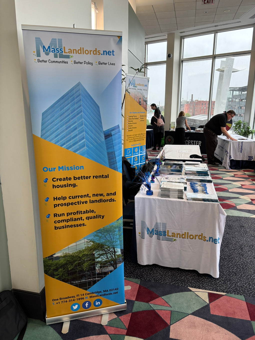 Two banners show MassLandlords mission and member benefits. A tablecloth shows the MassLandlords logo. The table is covered with pens, flyers and newsletters.
