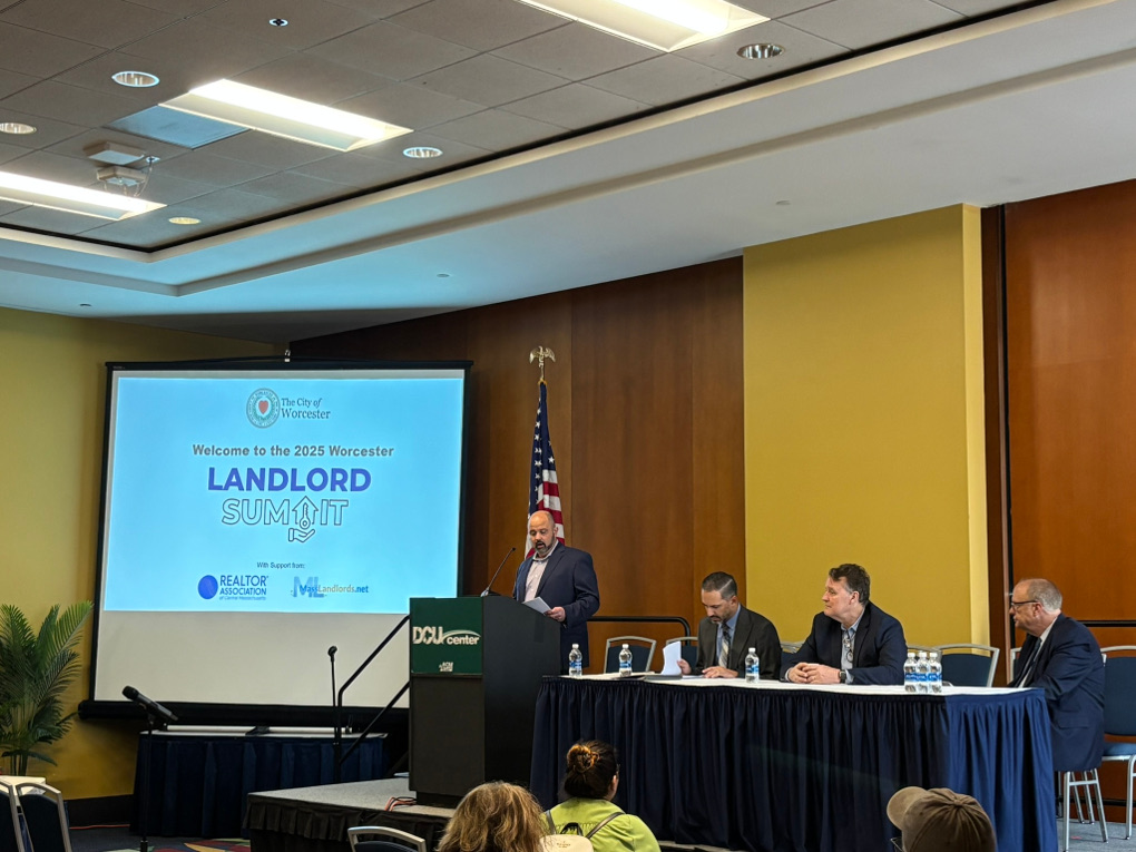 In a DCU center ballroom, Jim Brooks stands on a raised platform in a suit jacket at the podium. At the table to his left sit city manager Eric Batista, state senator Michael O. Moore, and mayor Joseph Petty. The title slide in the background reads, ‘Welcome to the 2025 Worcester Landlord Summit. Realtor Association of Central Massachusetts. MassLandlords.”