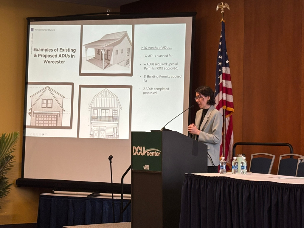 Michelle Smith stands at the DCU center podium in the ballroom. Her slide gives examples of proposed and existing ADUs in Worcester. It reads, ‘In 16 months of ADUs, 32 ADUs planned for, 4 ADUs required special permits (100% approved), 31 building permits applied for, 2 ADUs completed (occupied).’