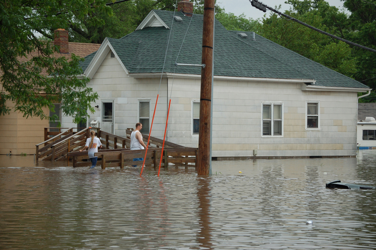 photo of a dirty white asbestos tile-sided home amid a deep pool of flood water, with a man and woman walking on a back deck, stairway and entrance ramp, also under water. 