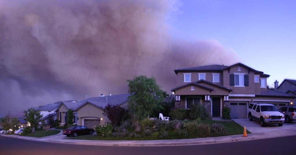 photo of a modern house alongside other houses in a suburban neighborhood with a massive smoke cloud looming behind them and filling the evening sky. 