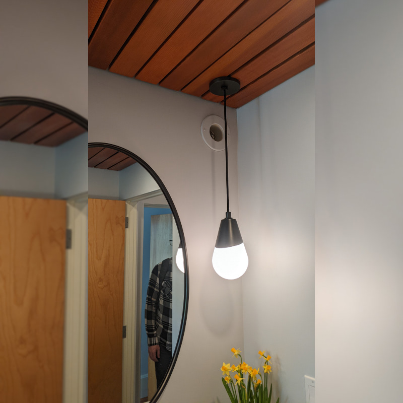 A bathroom mirror attached to the wall sits below and left of a mini-duct exit. A pendant light hangs in front of the duct.
