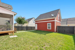 A backyard on a sunny day. A corner of a white house with back porch is visible on the left. On the right of the back yard sits a dark orange tiny home or renovated shed with door and windows.
