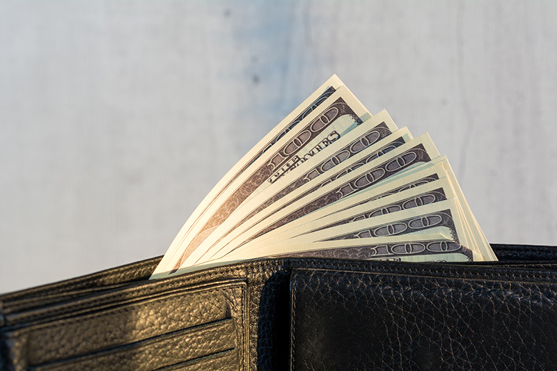 A black leather wallet is open against a pale blue background. Multiple hundred dollar bills are peeking over the edge of the section of the wallet designed to hold cash.