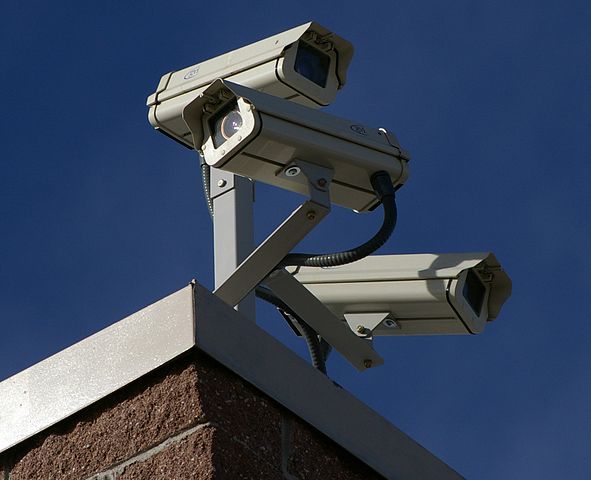 Three long focal length security cameras sit atop a brick building looking in different directions.