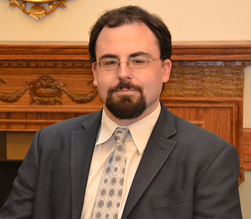Professional headshot of attorney Adam Sherwin, in dark gray suit and patterned tie, in front of a wood mantel. Attorney Sherwin wears glasses and a goatee and moustache. Board of Bar Overseers (editorial use).