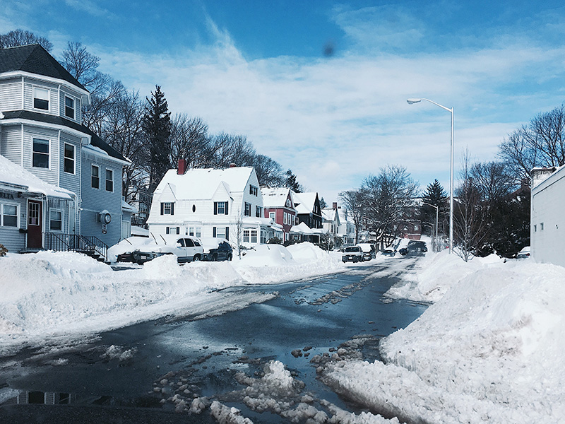 This image is a photograph of a plowed residential street in the wintertime, with high snow banks on either side of the road and slush around the banks. Houses are covered in snow, and cars parked on the street are blocked in with plowed snow.