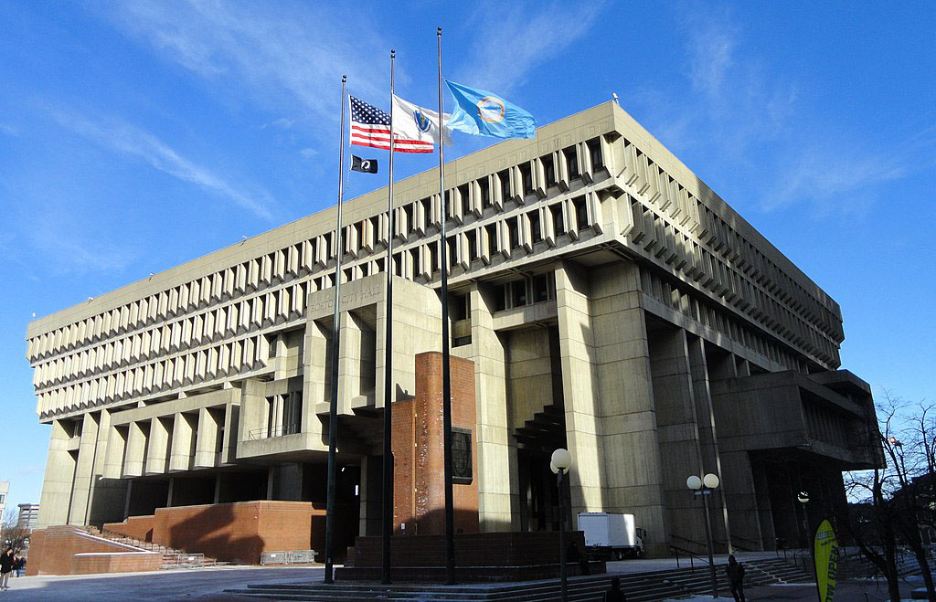 a photo of the exterior of Boston City Hall, a large, modern concrete-and-brick building, shown with the American and state flags flying in the wind.