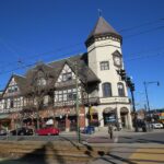 photo of Coolidge Corner, a thriving commercial intersection in Brookline, picturing a block with tudor castle-like architecture and ground-floor shops like Walgreens and others, pedestrians crossing the street and several cars parked along the curb.