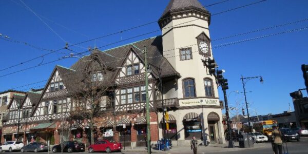 photo of Coolidge Corner, a thriving commercial intersection in Brookline, picturing a block with tudor castle-like architecture and ground-floor shops like Walgreens and others, pedestrians crossing the street and several cars parked along the curb.