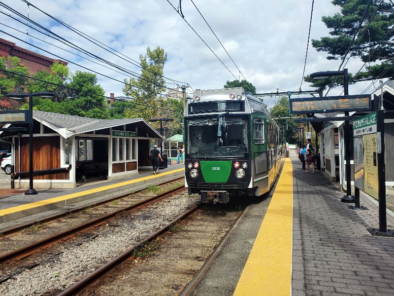 Photo of an incoming T train at Brookline Village stop with a platform showing train schedule information and people waiting to board, two train tracks running through, and a small, wooden station house.