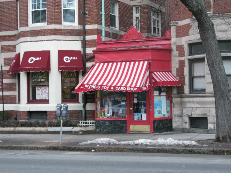 a photo of buildings on Brookline’s Harvard Street, focusing on a small shop called Irving’s Toy and Card Shop, sandwiched between two larger brick buildings with businesses and apartments.