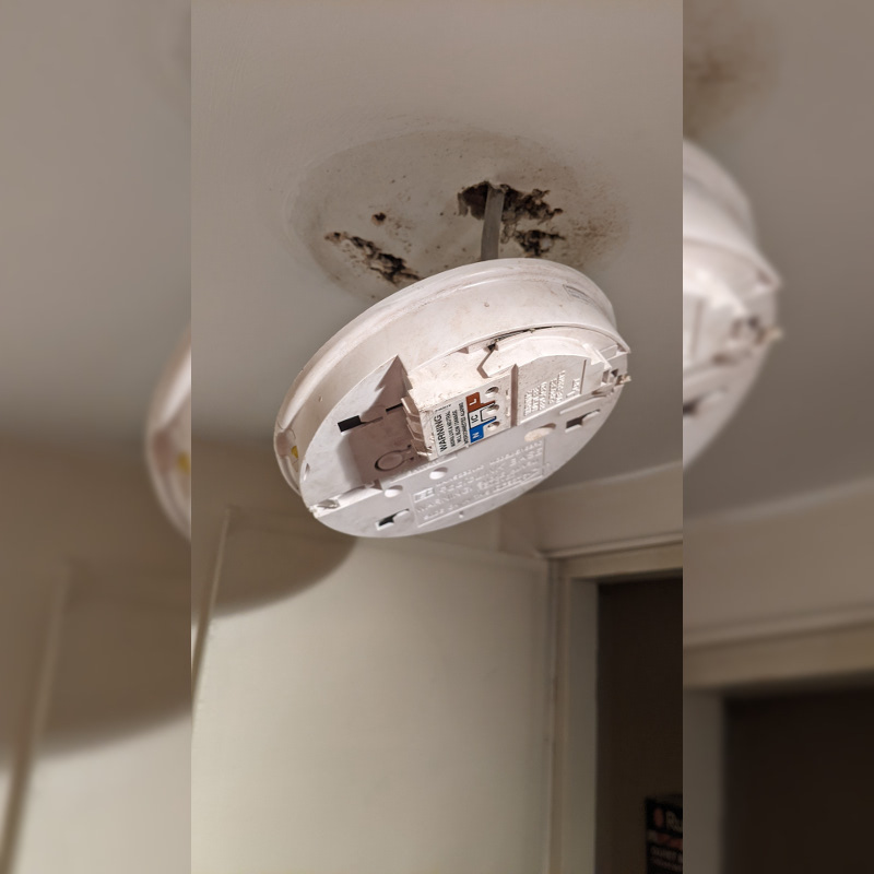 A UK-style smoke detector base hangs from a ceiling by a live wire. There is no detector on the base.