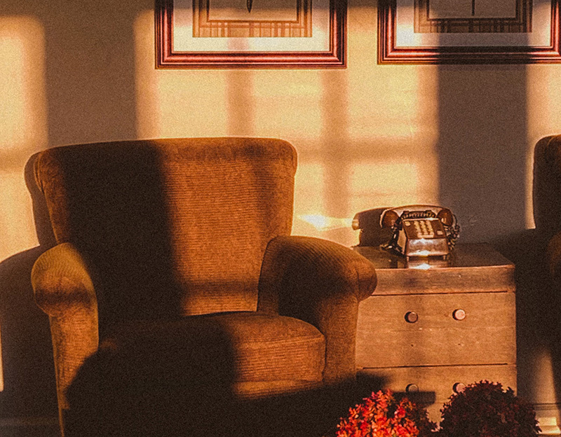 A corner of an empty apartment living room in late afternoon with sun coming in. The lower part of two photo frames on a beige wall are visible. An empty brown armchair sits in the corner. There is a brown coffee table and wood side table with a 1960s-style telephone sitting on top of it.