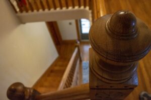 Closeup photo of a wooden staircase newel, with hardwood floor in the background and a staircase leading down to an exterior door.