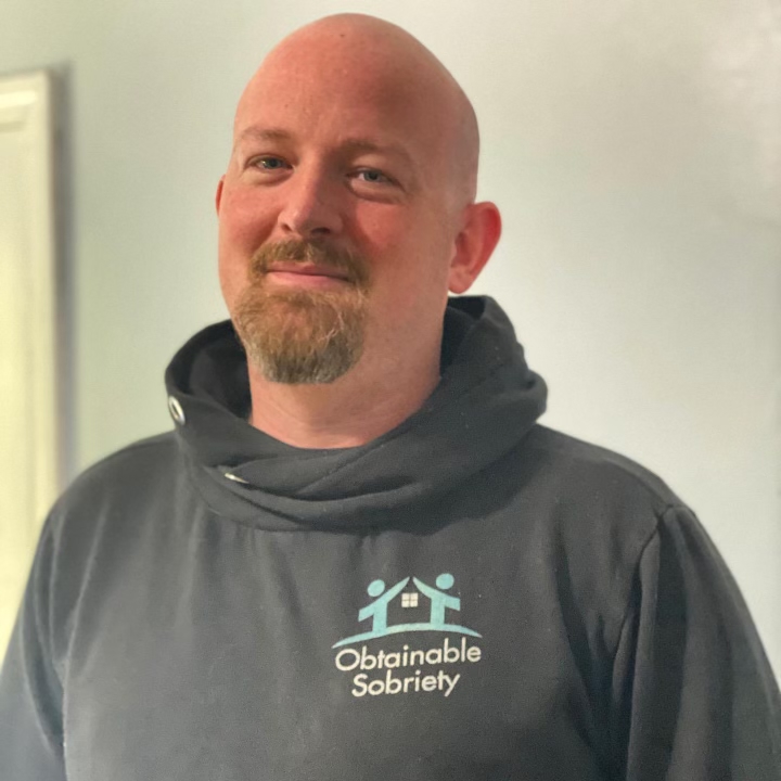 Picture of Patrick Sullivan. Patrick is wearing a gray hoodie with the logo of "Obtainable Sobriety" on it, two people with outstretched arms to form the roof of a house. Patrick wears a goatee and moustache. Image courtesy Patrick Sullivan.