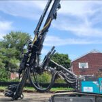 Photo of a large borehole drill, with tractor belt tires and blue-painted housing, with a brick house and multifamily building in the background, on a sunny day with wispy clouds.