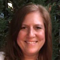 Sue McMahon is smiling fully. Her hair is shoulder length and parted to the side. She is standing outdoors in front of a tree.