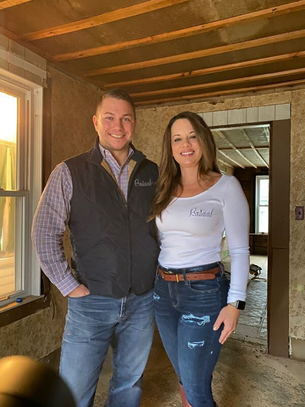 A husband and wife, Matt and Kelli Slowik, stand facing the camera in a room of a house they are renovating, with open beams on the ceiling and walls.