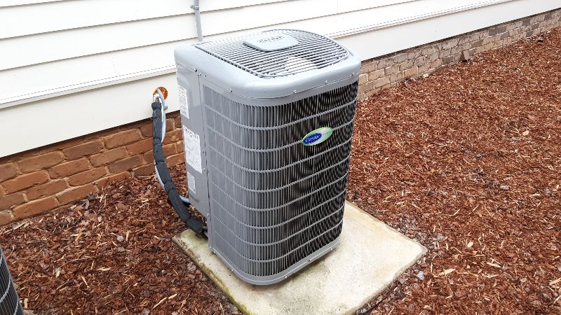 A close up photo of a heat pump exterior unit sitting in mulch next to the outside wall of a house, showing part of a brick foundation and house siding.