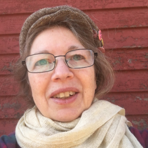 close up headshot photograph of Jo Landers, who has brown hair and wears glasses. She wears a brown newsboy cap, white scarf, and purple and green plaid flannel shirt. She is smiling fully.