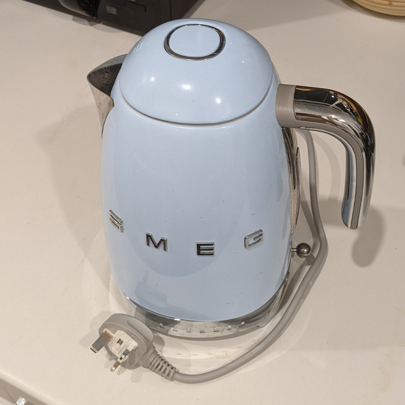 A round metal tea kittle with short cord and chunky plug sits on its base. The letters “SMEG” are embossed on its side.