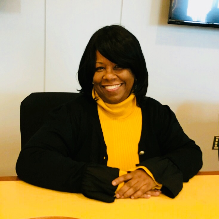 Picture of Val Mayo smiling wearing a dark jacket and light turtleneck reminiscent of the color pattern of of a bee.