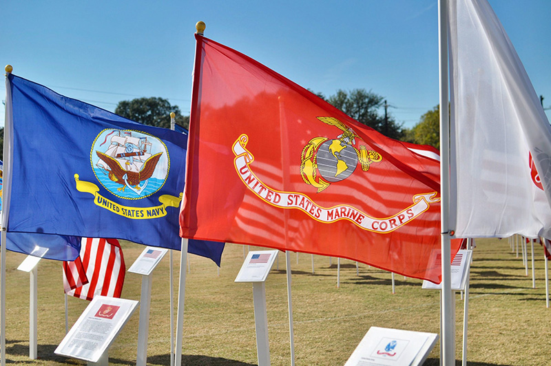 Three flags wave in front of a grassy field. A blue one is for the U.S. Navy, a red one is for the U.S. Marine Corps. The third, a white flag, does not have a clear insignia.