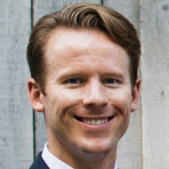 Matthew Pelrine smiling and wearing a suit and tie in front of a wood clapboard background.